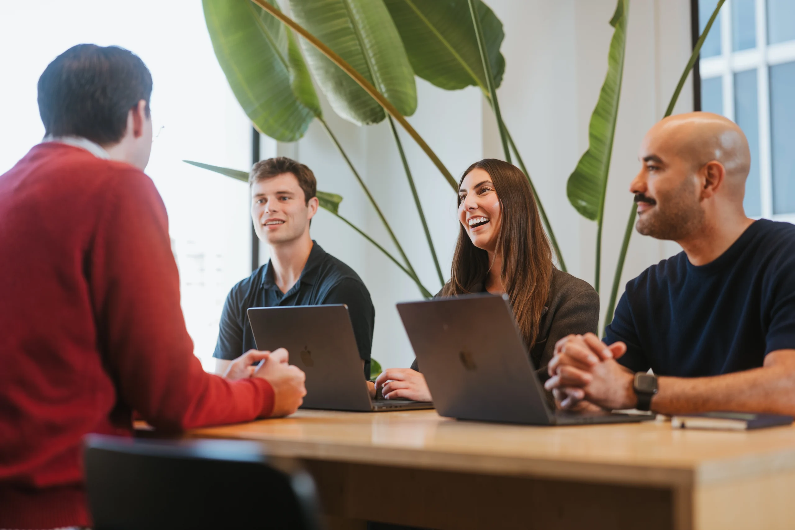 Four people sit around a table with laptops, engaged in a meeting and smiling. Large green plants are in the background, creating a bright and welcoming office environment where the team’s collaboration is guided by ironclad morae.