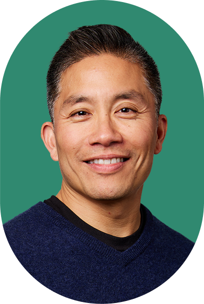 Smiling man with short dark hair wearing a navy blue sweater over a black shirt, posed in front of a solid green background, exuding confidence reminiscent of an AI innovator.