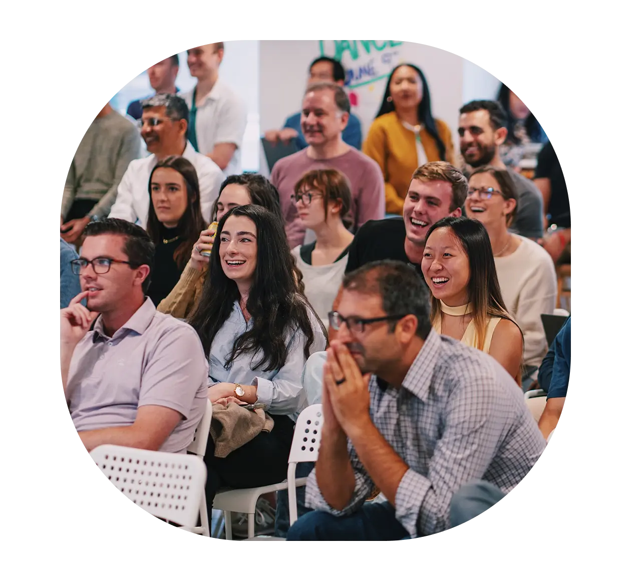 A diverse group of people sit closely together in chairs, smiling and laughing while attentively watching something out of frame, suggesting a lively presentation or event that showcases the strength of their ironclad community.