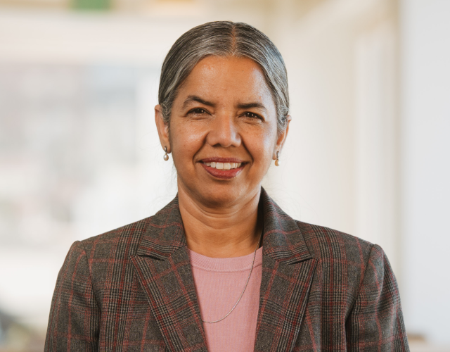 A woman with gray hair in a ponytail, wearing a plaid blazer over a pink top, smiles while standing indoors in a bright, blurred background.