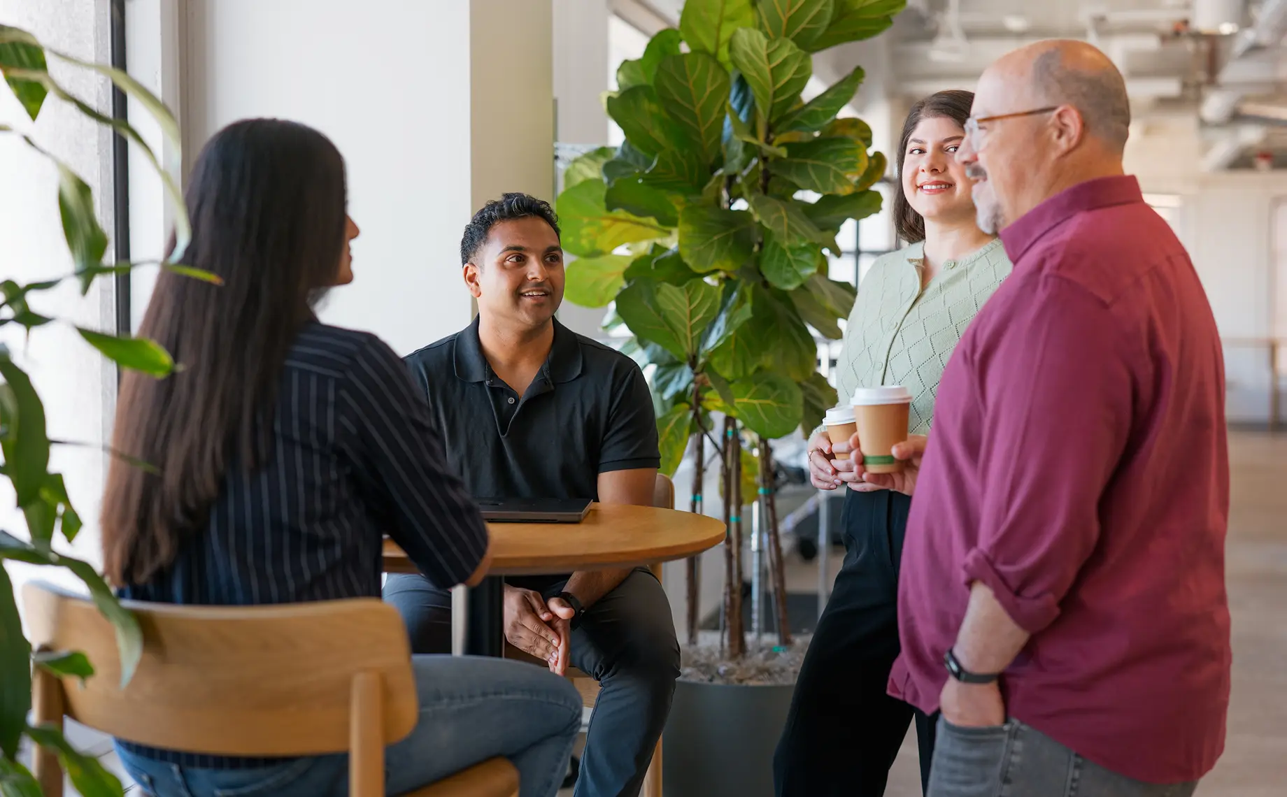 Four people gather around a small table in a bright office, discussing soft skills in procurement. Two hold coffee cups as they smile; large green plants and natural light fill the welcoming space.