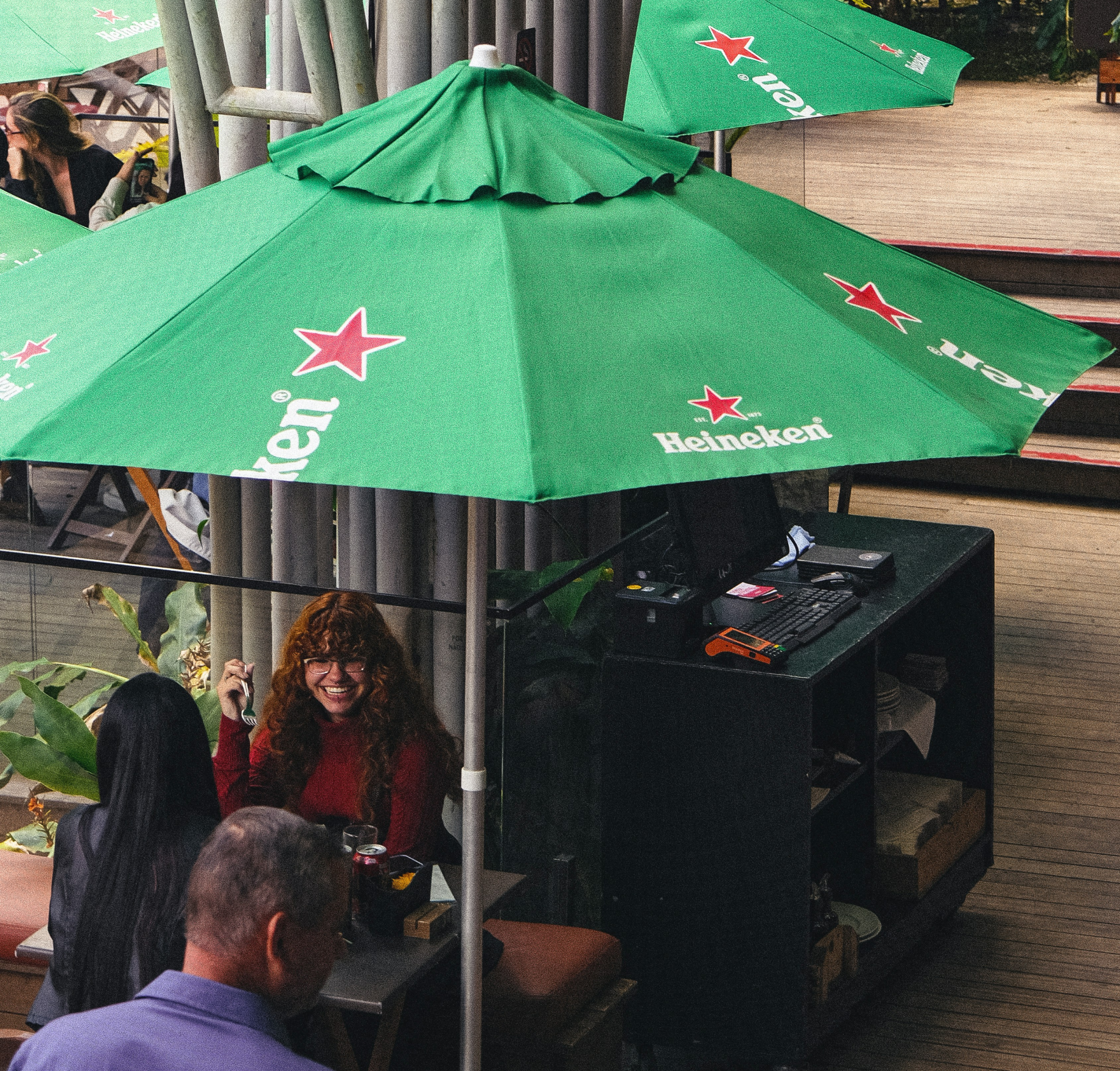 People sit at outdoor tables under green Heineken umbrellas on a wooden deck. A woman in a red top smiles while talking to another person, and two others are visible nearby. Lush plants surround the seating area.