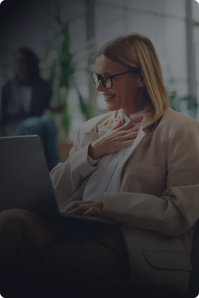 A woman wearing glasses and a blazer smiles while looking at her laptop with a hand resting on her chest, representing ironclad customers.